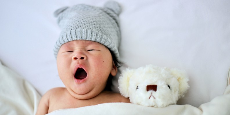 Gabende baby ligger i sengen med hue på og bamse i favnen